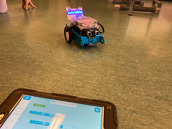 mBot -robotti lattialla. Etualalla iPad, jossa mBlock Blockly-sovellus